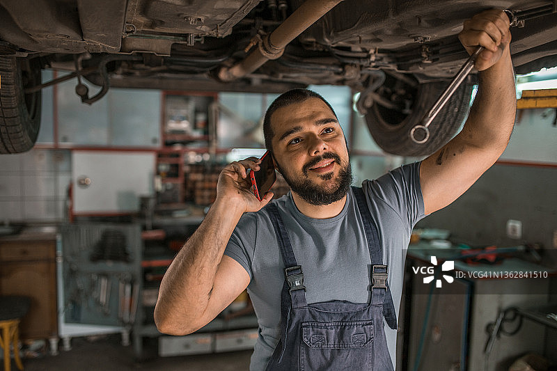 在汽车修理店，汽车修理工微笑着打电话修理汽车图片素材