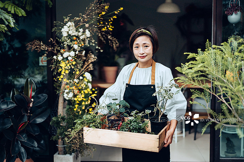自信的亚洲女性花店的肖像，小生意花店的老板。在她的花店外，她拿着一个装满鲜花和植物的木箱。微笑着看着镜头。喜欢和花在一起的工作。中小企业的概念图片素材