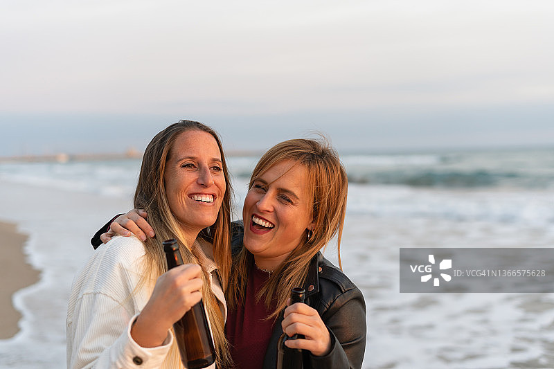 快乐的女性朋友喝着啤酒，在日落时分的海滩上玩耍图片素材