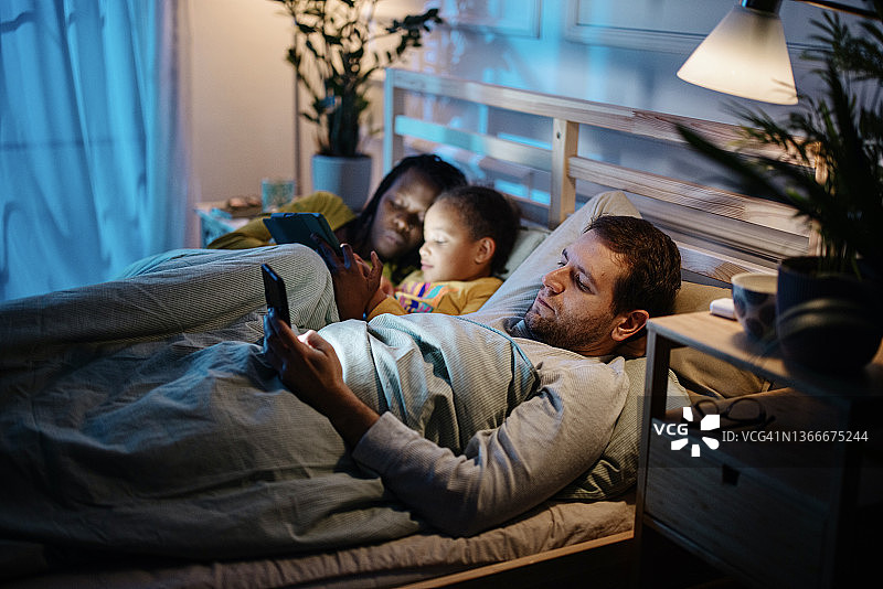 母女俩睡前在平板电脑上读故事，而爸爸则在智能手机上看书图片素材