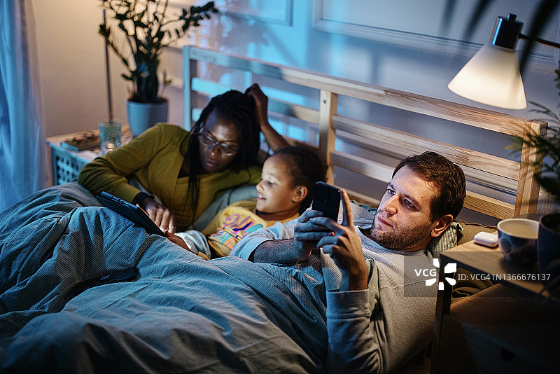 母女俩睡前在平板电脑上读故事，而爸爸则在智能手机上看书图片素材