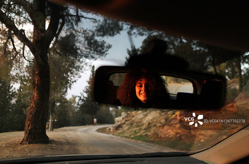 年轻女子在夕阳下对着汽车的后视镜微笑，汽车停在森林里图片素材