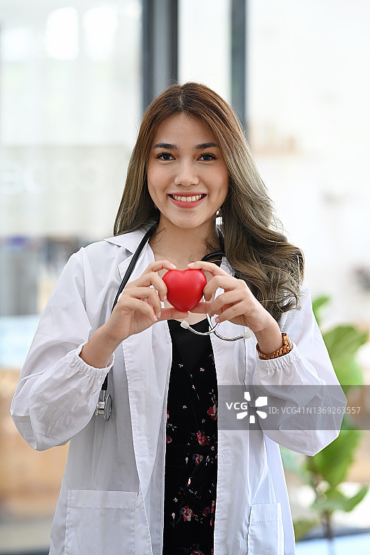 身穿白色制服的女医生拿着红心，对着镜头微笑。医疗保健和心脏病学概念。图片素材