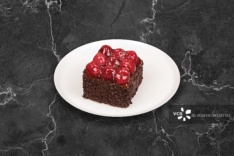 树莓巧克力蛋糕，方形。它被盛在一个白色的盘子里，背景是黑色的。图片素材