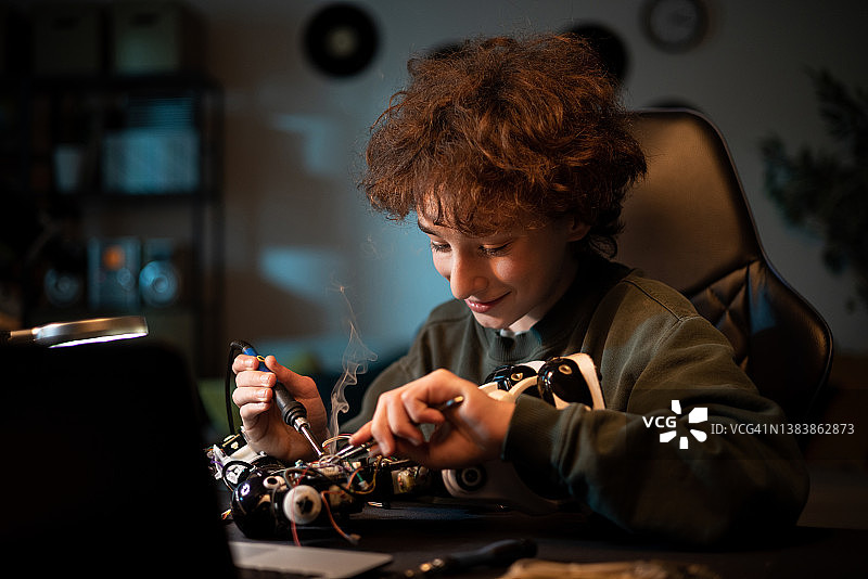 一个微笑的男孩正在学习电子技术，焊接机器人领域的电线和电路板。一个孩子深夜在房间里摆弄玩具图片素材