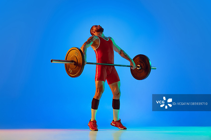 工作室拍摄的年轻人在红色运动服锻炼与杠铃孤立的蓝色背景霓虹灯。运动，举重，力量，成就概念图片素材