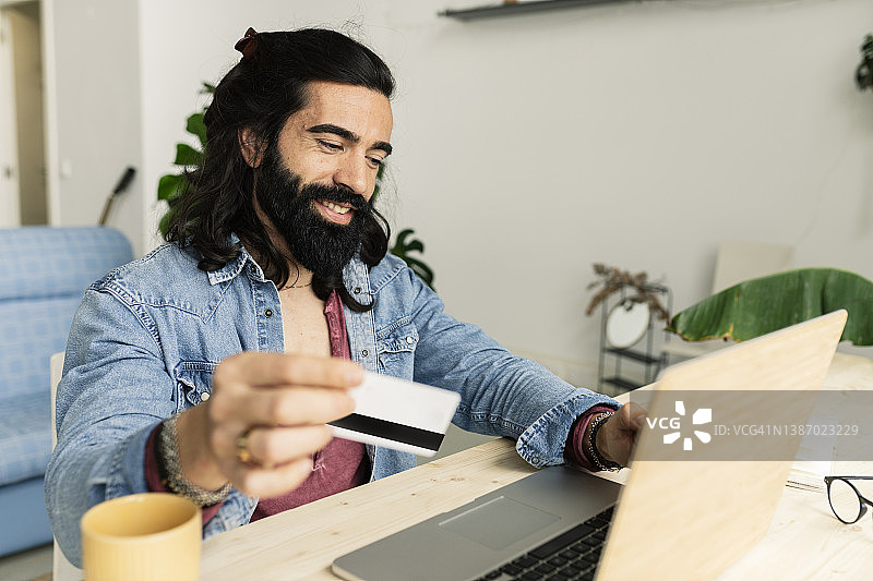 英俊，大胡子，长发的男人在家用信用卡网购图片素材