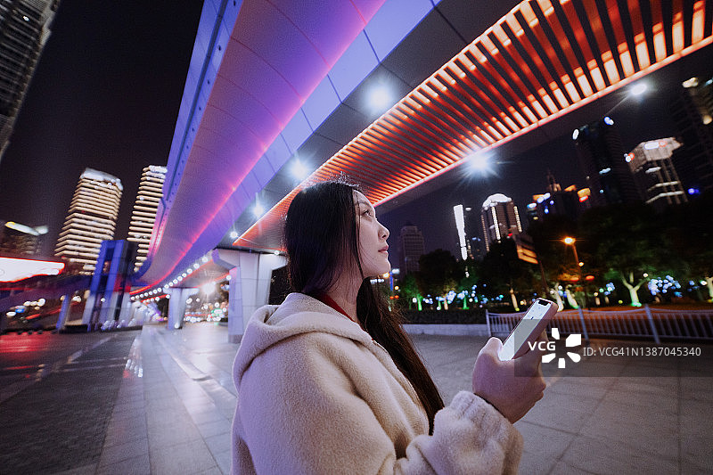 一名年轻的亚洲女子站在夜间发光的行人天桥下使用手机图片素材