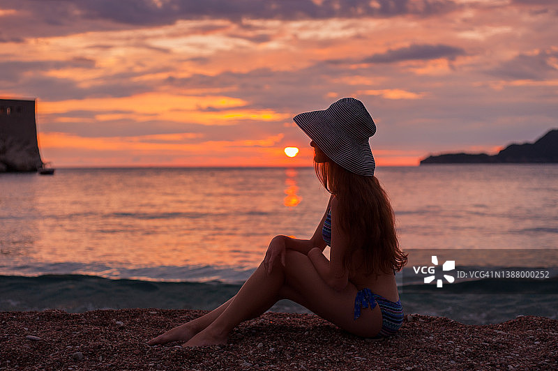 女人坐在太阳帽上享受在海滩上对着天空的彩色日落图片素材