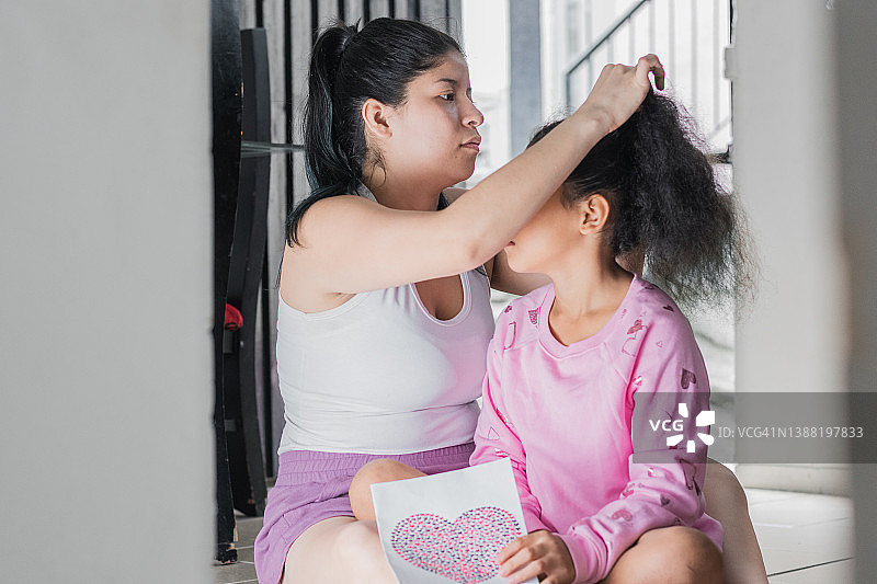 美丽的拉丁母亲与她的黑发女儿，梳理她的头发或使马尾辫坐在她的房子的地板上。年轻的母亲和女儿说话。家庭的概念。图片素材