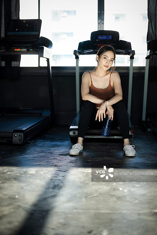 在健身房的穿线跑步机上锻炼的年轻亚洲女性的肖像。快乐的肌肉健康的女性在健身中心锻炼，通过在线健身教练使用笔记本电脑进行训练图片素材
