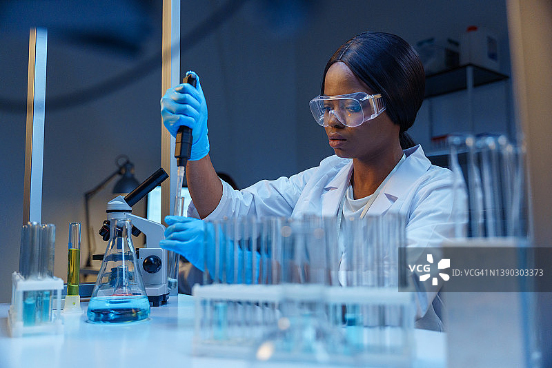 一个年轻美丽的微生物学家，在现代化的实验室里，戴着防护眼镜，在她的桌子上用微型吸管工作，在试管架上用实验室烧瓶和玻璃试管图片素材