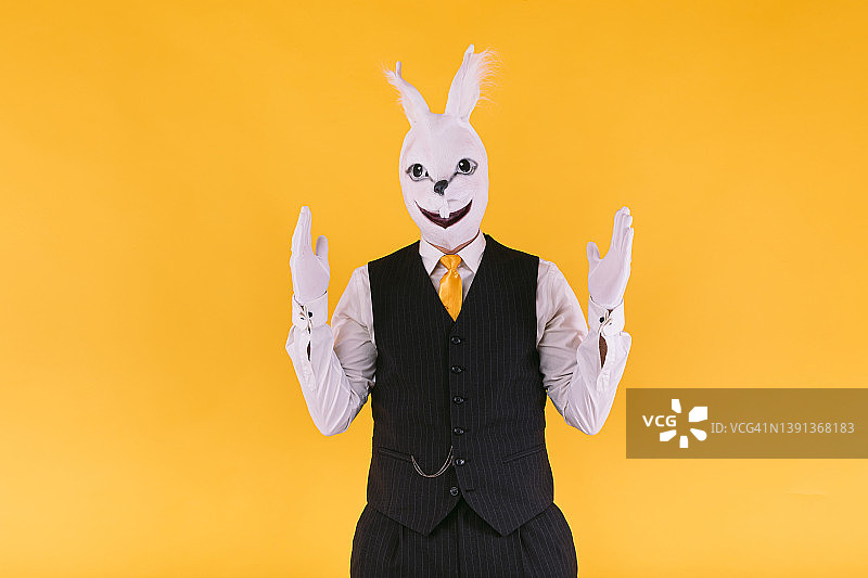 变相的人戴着兔子面具，穿着西装外套，背心和领带，举起双手，在黄色的背景上。嘉年华，派对，复活节和庆祝的概念。图片素材