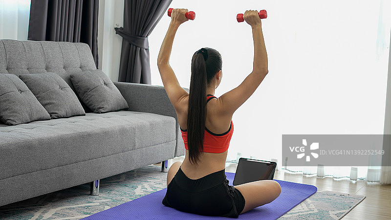年轻的女运动员在瑜伽垫上用平板电脑做哑铃练习图片素材