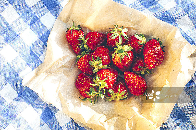 牛皮纸上有新鲜的甜草莓，蓝色野餐毯上有方格图案。夏季野餐的概念图片素材