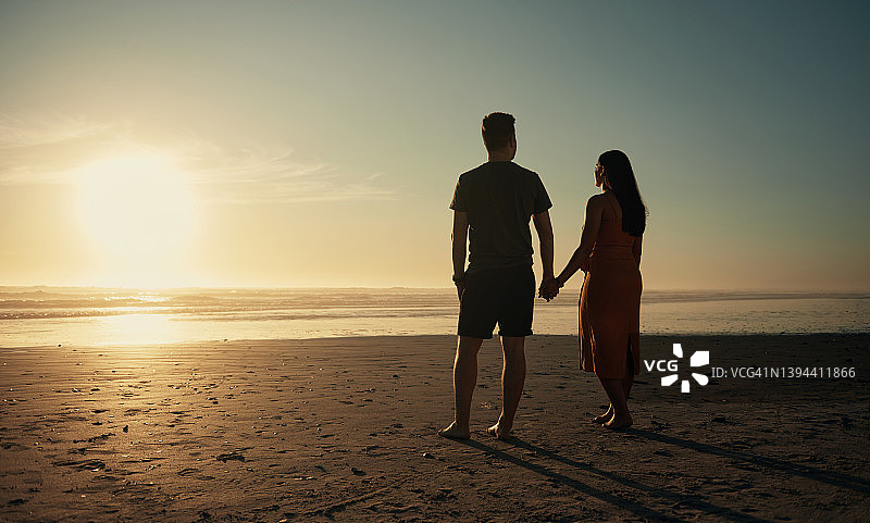 后视图两个年轻夫妇牵着手站在海滩上看日落日出度假的一天图片素材