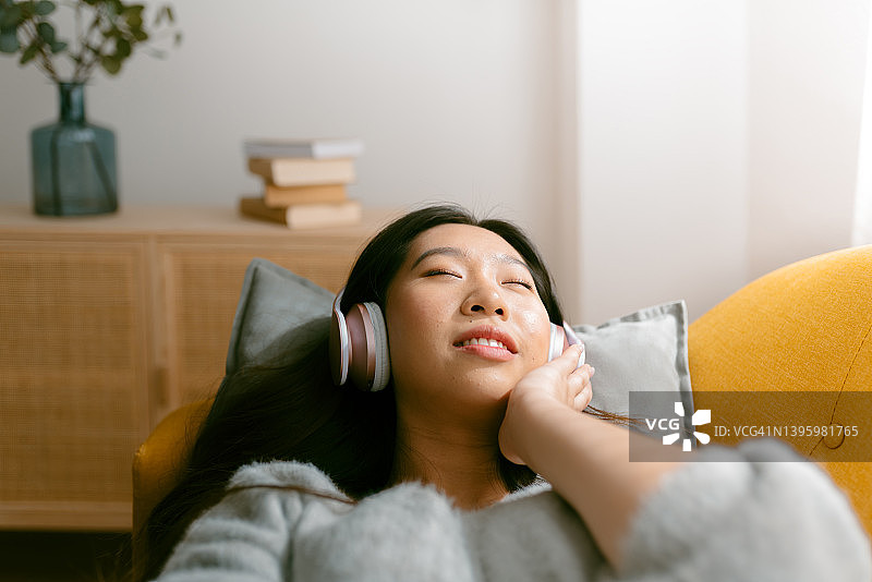 一个年轻的亚洲女人听音乐与耳机放松地躺在家里的沙发上的前视图。图片素材