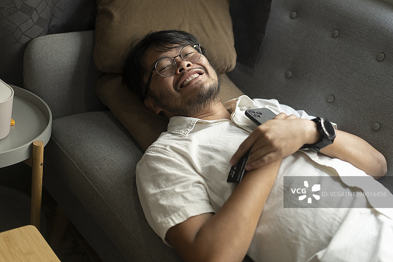 积极的情绪亚洲年轻人躺在沙发上和幸福后，网上购物与批准信用卡在家里。图片素材