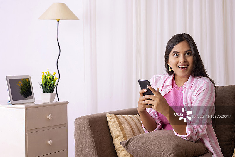 美丽的印度女孩坐在沙发上用智能手机摆姿势图片素材
