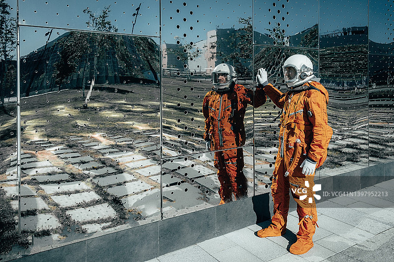 身穿橙色太空服、头戴太空头盔的宇航员站在一面巨大的镜子旁图片素材