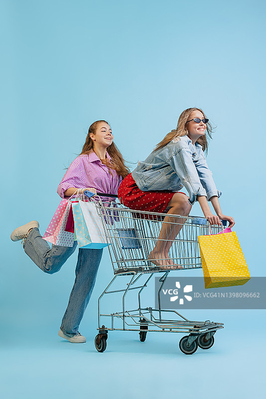 工作室拍摄的漂亮快乐的女孩与购物袋骑在一辆车孤立的蓝色背景。销售概念，黑色星期五，折扣，情感，青春和时尚图片素材