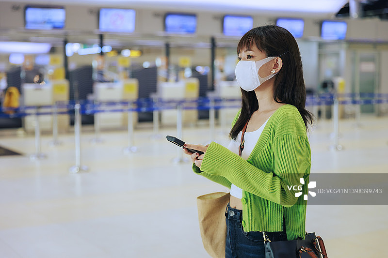 年轻的亚洲女企业家戴着口罩站在机场出境区，用手机阅读电子邮件。商务旅行的概念。图片素材