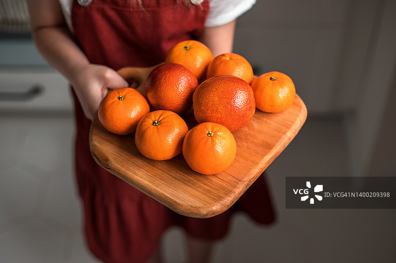 孩子的手拿着砧板和柑橘在家。快乐的小女孩带着水果在家里。图片素材