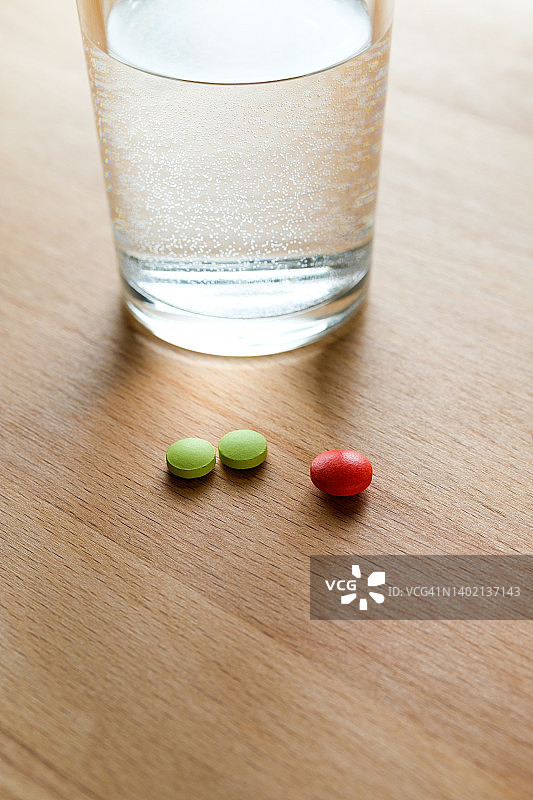 厨房的木桌上放着红色和绿色的药丸和一杯水。服用维生素、止痛药、药物和膳食补充剂。疾病预防和治疗的概念，保健和医药。为怀孕做计划和准备。图片素材