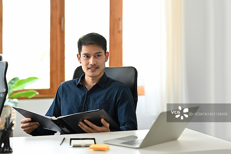 一名年轻的亚洲男子坐在办公室里的画像，自信地看着镜头，手里拿着一份报告、数据和文件，为商业和技术概念。图片素材