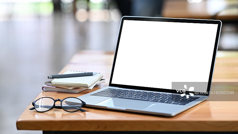 白色空白显示屏的笔记本电脑，木制办公桌上的眼镜和笔记本。图片素材