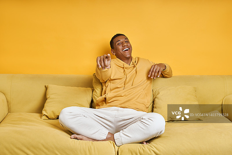快乐的混血男子坐在黄色背景的沙发上指着摄像机图片素材