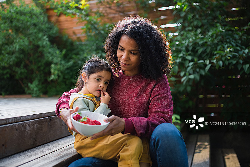 多种族妇女和她的女儿坐在花园里，吃着草莓享受夏天的时光。图片素材