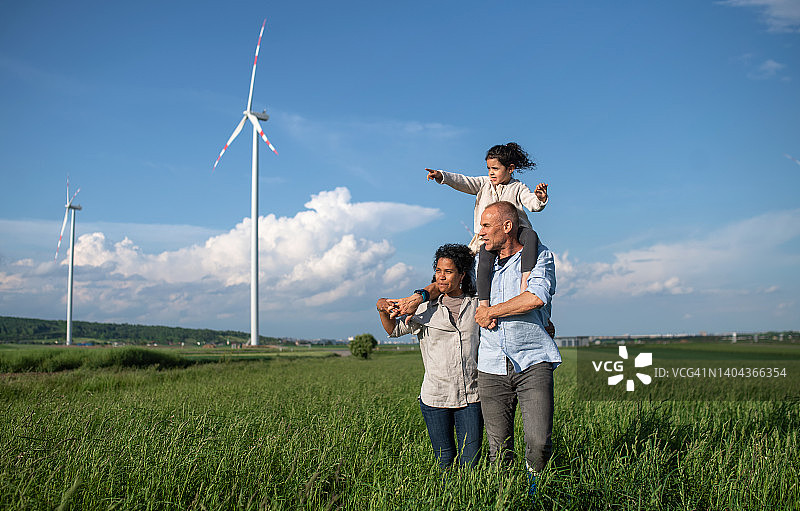 混血家庭微笑着走在田野里。风力发电机的背景，可再生能源的概念。图片素材