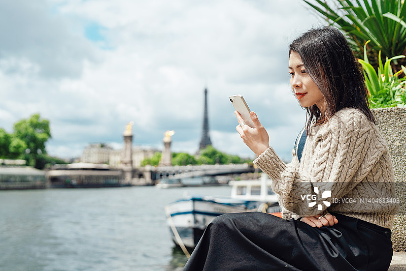 年轻的亚洲女性坐在塞纳河边使用智能手机图片素材