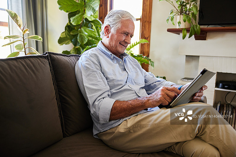 一位面带微笑的老人一边在沙发上放松，一边使用平板电脑图片素材