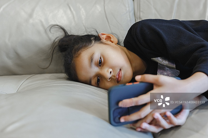 小女孩躺下玩看智能手机在线学习。小女孩在线聊天学习。屏幕上的小女孩，社交媒体成瘾。图片素材
