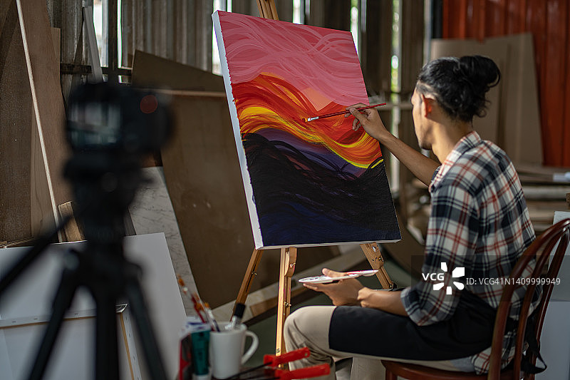 年轻的亚洲男艺术家，他坐在大画板前并在线直播绘画课。一名亚洲男子周末早上在家里的画室画画图片素材