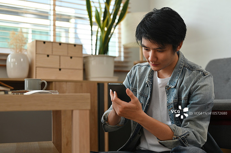 与亚洲帅哥在客厅使用智能手机的肖像，家庭生活理念。图片素材