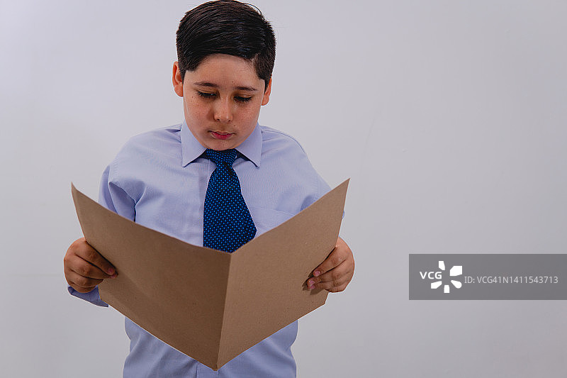 一个男孩穿着西装打着领带假装自己是一个商人，同时阅读着一个重要文件的文件夹。小男孩打扮成商人。图片素材