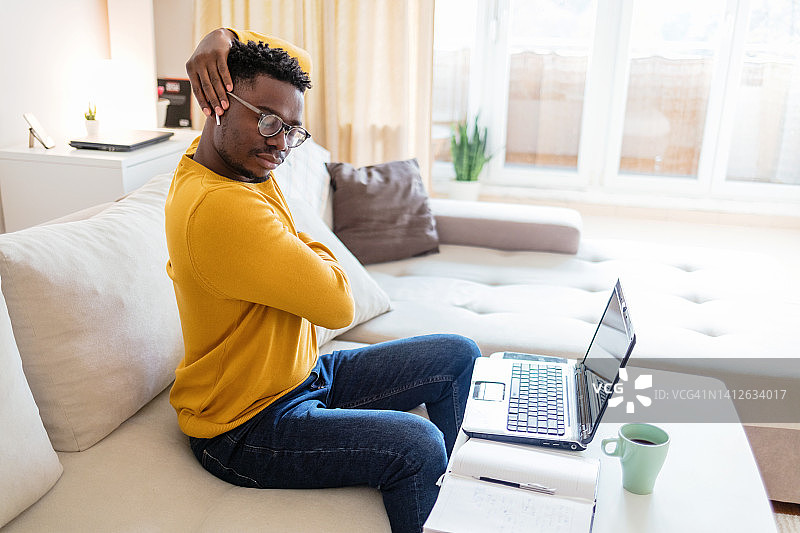 疲惫的非洲商人在家对着电脑工作，感到肩膀疼痛。男人坐在家里用笔记本电脑工作时肩膀和背部疼痛。图片素材