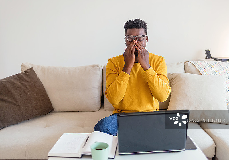 精疲力竭的非洲裔美国人办公室职员坐在办公桌前，拿着笔记本电脑，用手捂着脸，疲惫的人闭上了眼睛。这是一个年轻的商人在工作期间感到疲劳的照片。图片素材