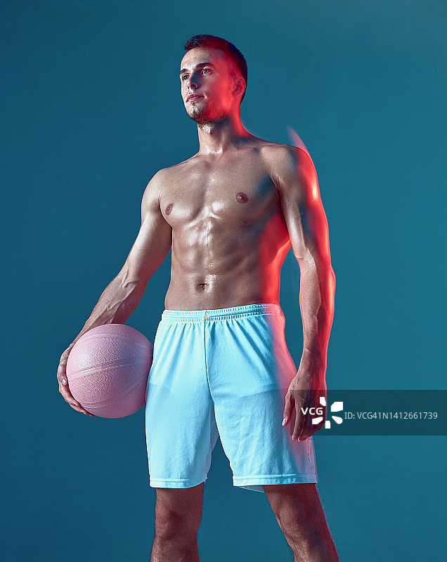 男性篮球运动员，在蓝色的演播室背景上，赤裸的肌肉躯干拿着球。长时间的曝光。体育锻炼图片素材