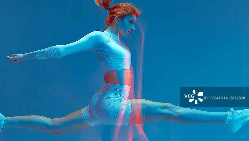 运动女孩在蓝色的背景上跳劈叉。动态模糊，长时间曝光。体操锻炼、健身、运动图片素材