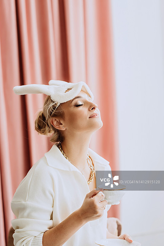 年轻美丽的女人在一个春天复活节兔子的白色面具与一杯咖啡在她的手。狂欢节新年面具。软选择聚焦。图片素材