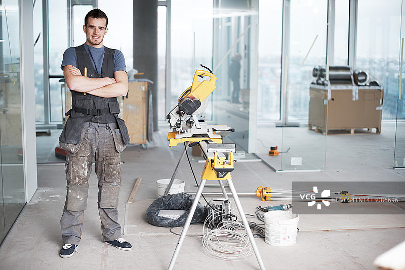 一个快乐的年轻承包商的肖像与双臂交叉站在建筑工地的工业建筑项目。工程师建筑工人使用电锯设备工具翻新建筑物图片素材