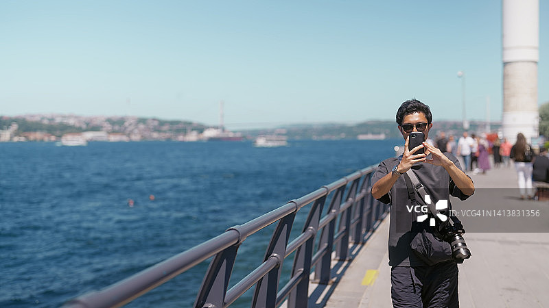 年轻的男性游客在海边散步，用手机拍摄视频和照片图片素材