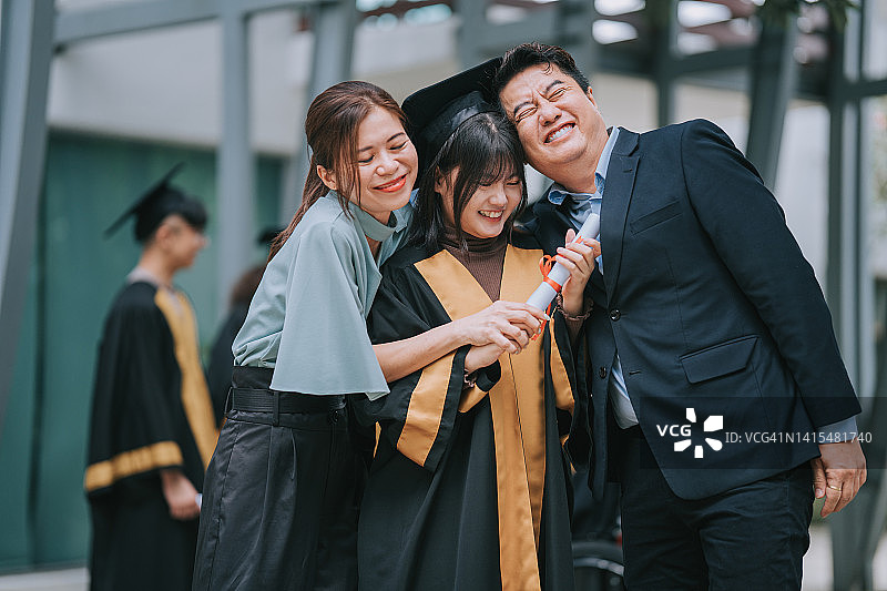 自豪的亚裔华裔父母带着穿着大学毕业礼服的女儿站在大学大楼外图片素材