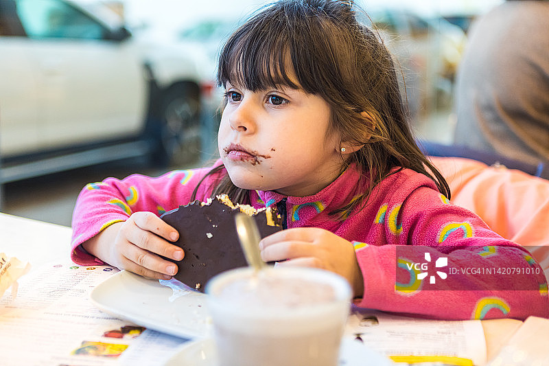 穿着粉色毛衣的小女孩在自助餐厅，吃着巧克力松饼，嘴巴和手都很脏。图片素材