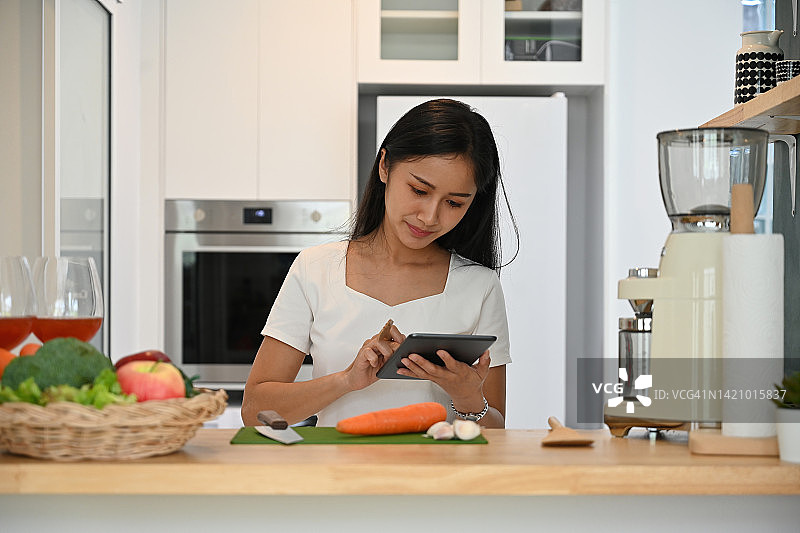 20多岁的年轻素食女性使用数字平板电脑寻找食谱或在家里的厨房里用蔬菜在线学习烹饪，家庭生活和健康的生活方式概念。图片素材