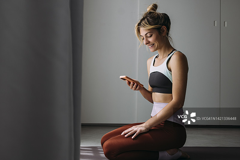 一个快乐的美丽的女人正在做瑜伽，使用她的手机应用程序，而坐在她的瑜伽垫上图片素材